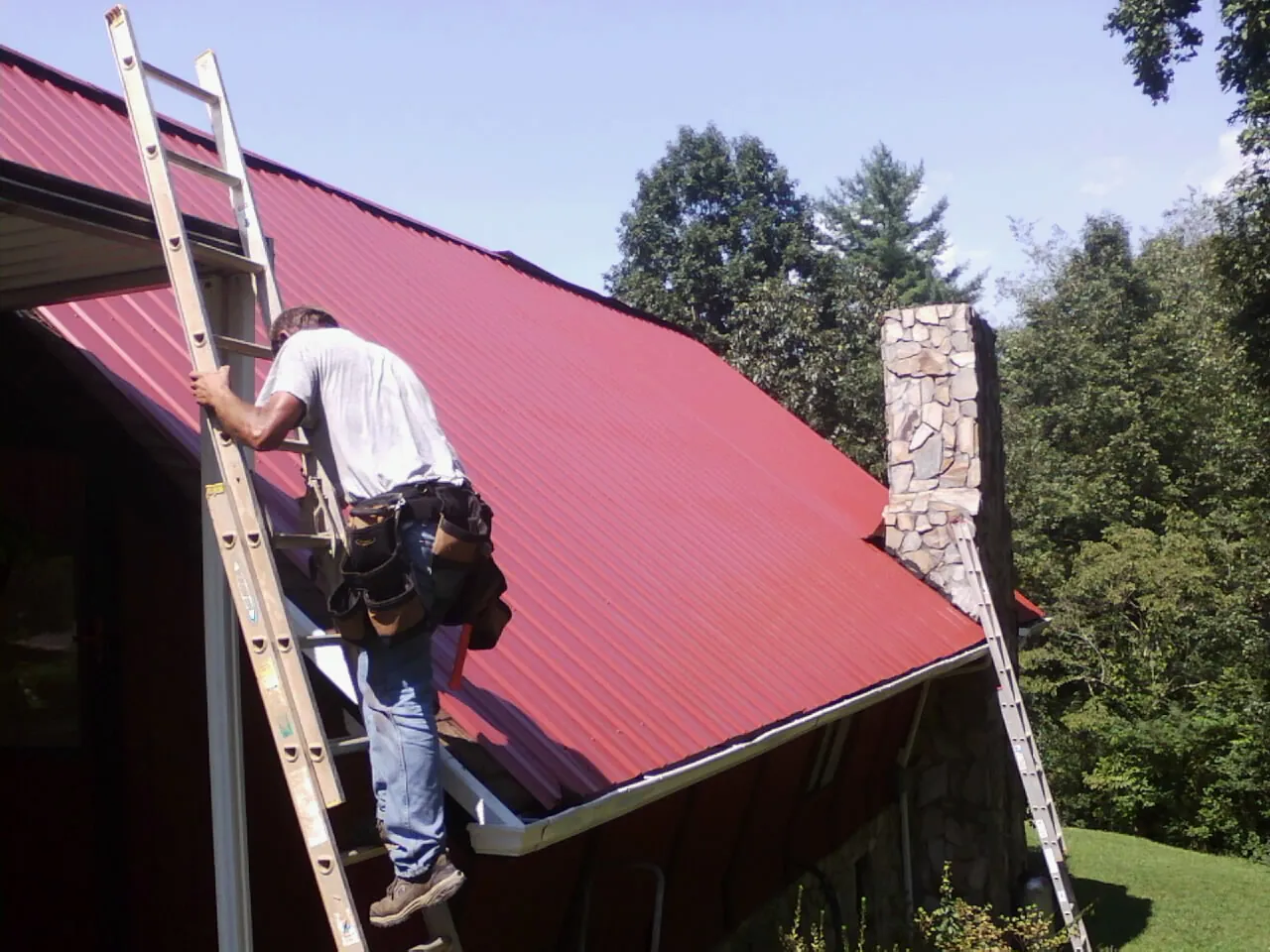 Roofer on a ladder beside a red metal roof and stone chimney — Soffit & Fascia Repair by Roofing Brothers in Budd Lake
